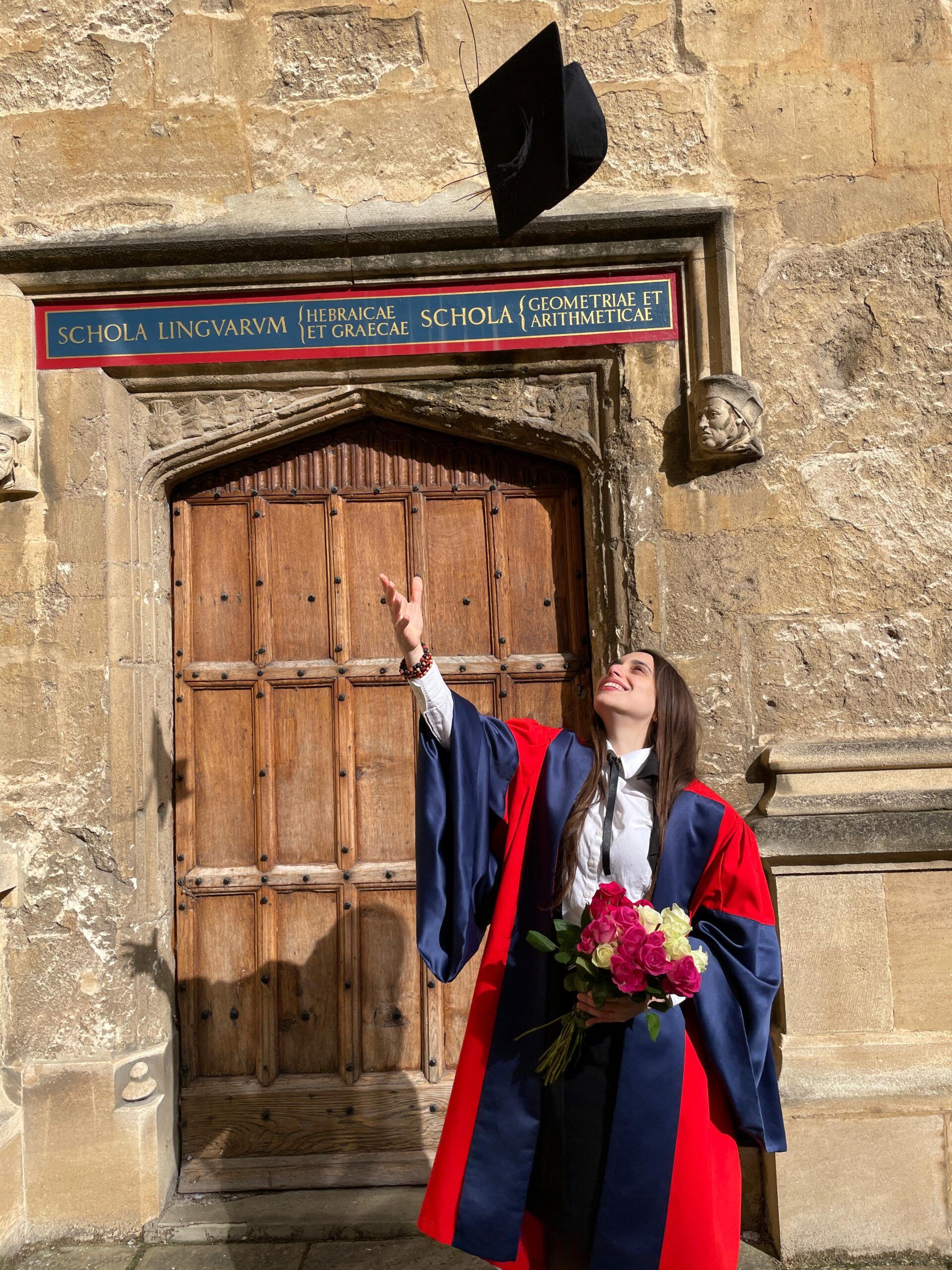 A graduate in academic dress holding flowers and tossing a cap into the air outside a stone doorway.