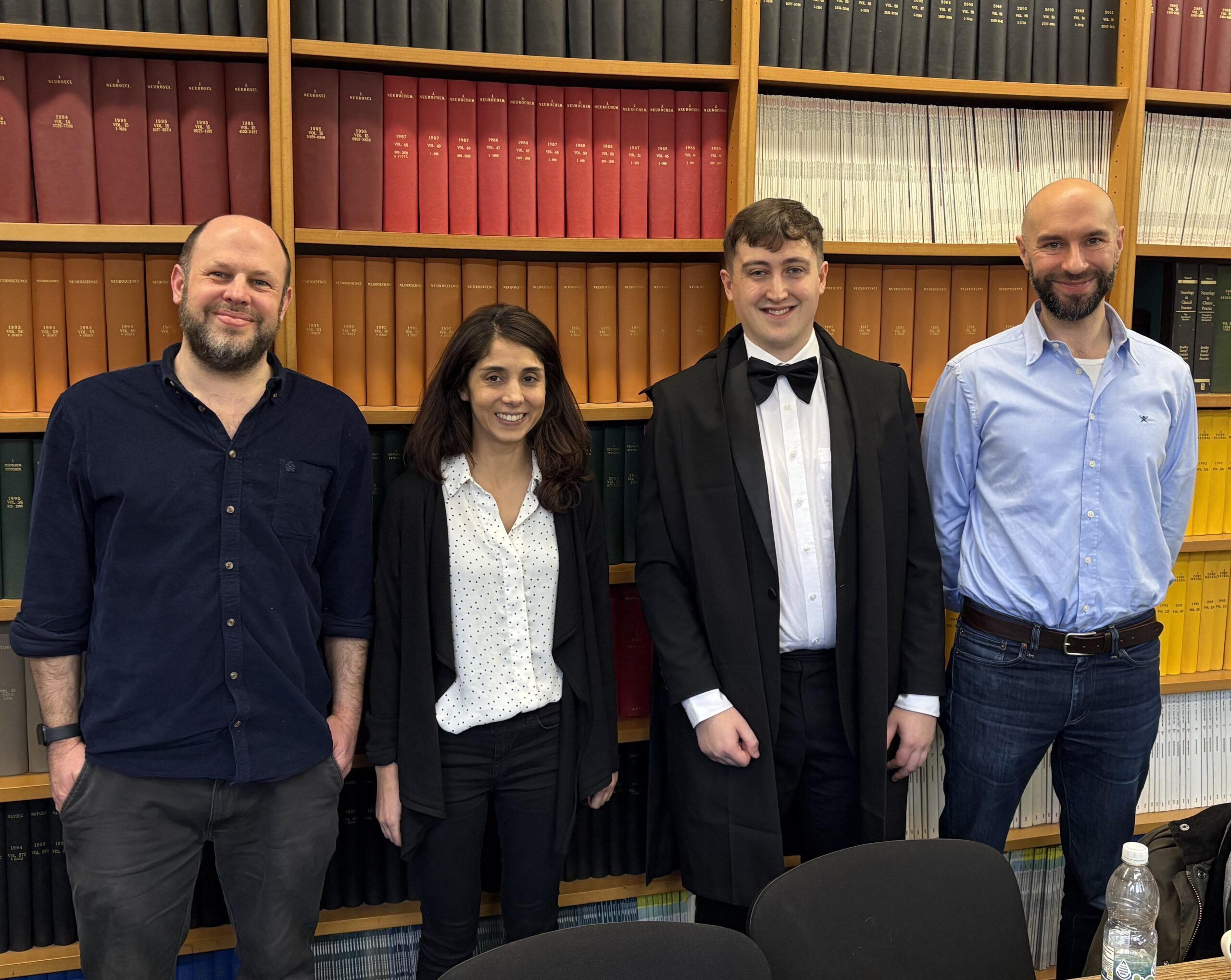 Four people standing in front of library bookshelves on the day of Isaac’s PhD viva.
